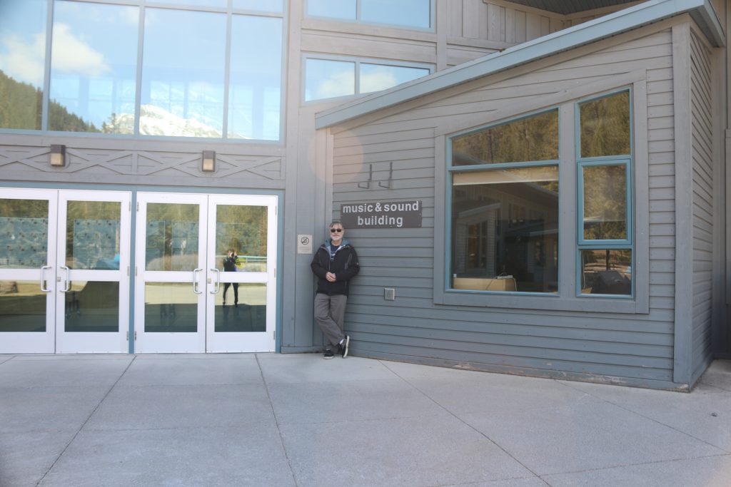 Picture of author standing in front of main door of the Banff Art Centre. the roof of the building slopes up toward the right and two stainless steel windowed doors can be seen to the left of the author who is wearing grey pants and a black jacket