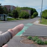 Image shows an intersection of a road with a green painted crosswalk and in the background a road can be seen sloping upwards. In the foreground there is a hand with a finger pointing up the hill. Trees can be seen lining the street.