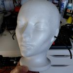 Image shows and white styrofoam female manequin head with wires sticking out of the sides where the ears are. In the background can be seen an assortement of things strewn on a workbench including small containers, toy cars, a Netscape mug with pens.