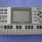 Picture shows a silver musical instrument with a large LCD screen in the middle and a row of keys along the bottom arranged like piano keys in two octaves. It has small buttons on the right and left of the screen with various functions for playing, recording and editing music.