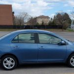 Image shows a blue four door Sedan with aluminium sport wheels parked in a parking lot space. Buildings can be seen the background