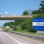 Image shows a view of a sign welcoming the motorist to the province of Ontario. The image shows the sign on the right hand side, there is an overpass just beyond the sign passing over the highway and greenery and grass can be seen along the highway behind a short fence and some trees beyond.