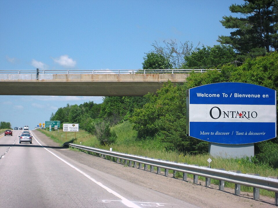 Image shows a view of a sign welcoming the motorist to the province of Ontario. The image shows the sign on the right hand side, there is an overpass just beyond the sign passing over the highway and greenery and grass can be seen along the highway behind a short fence and some trees beyond.